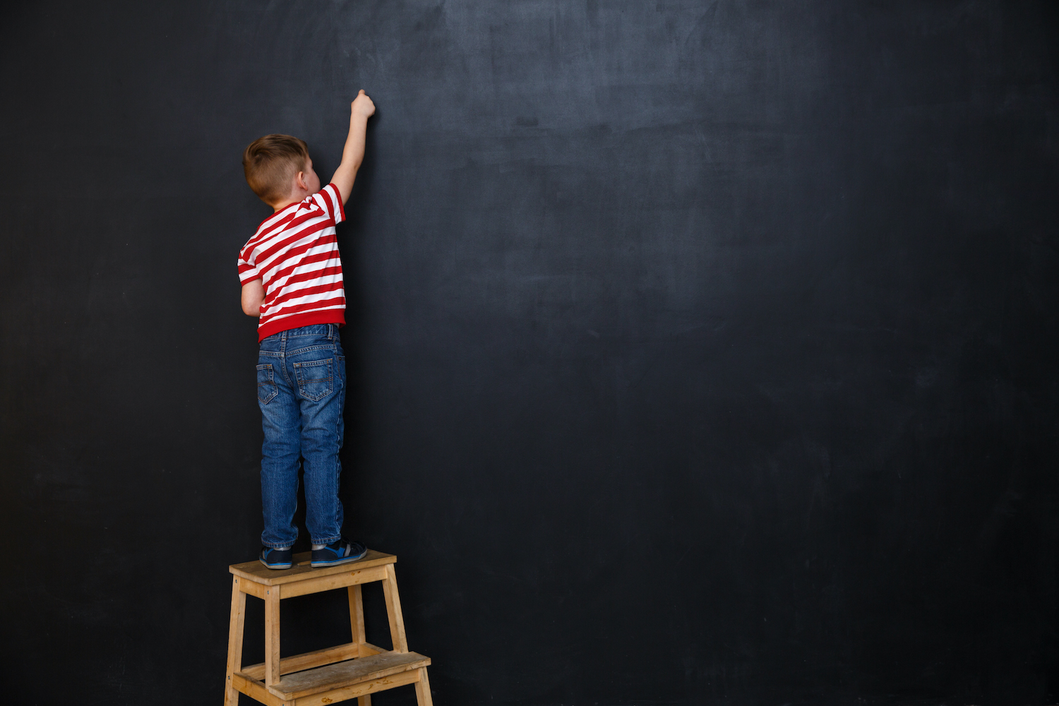 Back view of a little boy standing on ladder and writing with chalk on the backboard in school class